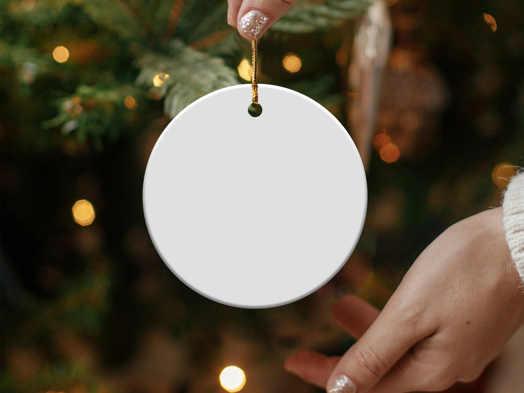 Blank white ceramic Christmas ornament held by a hand in front of a blurred tree.