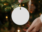 Blank white ceramic Christmas ornament held by a hand in front of a blurred tree.