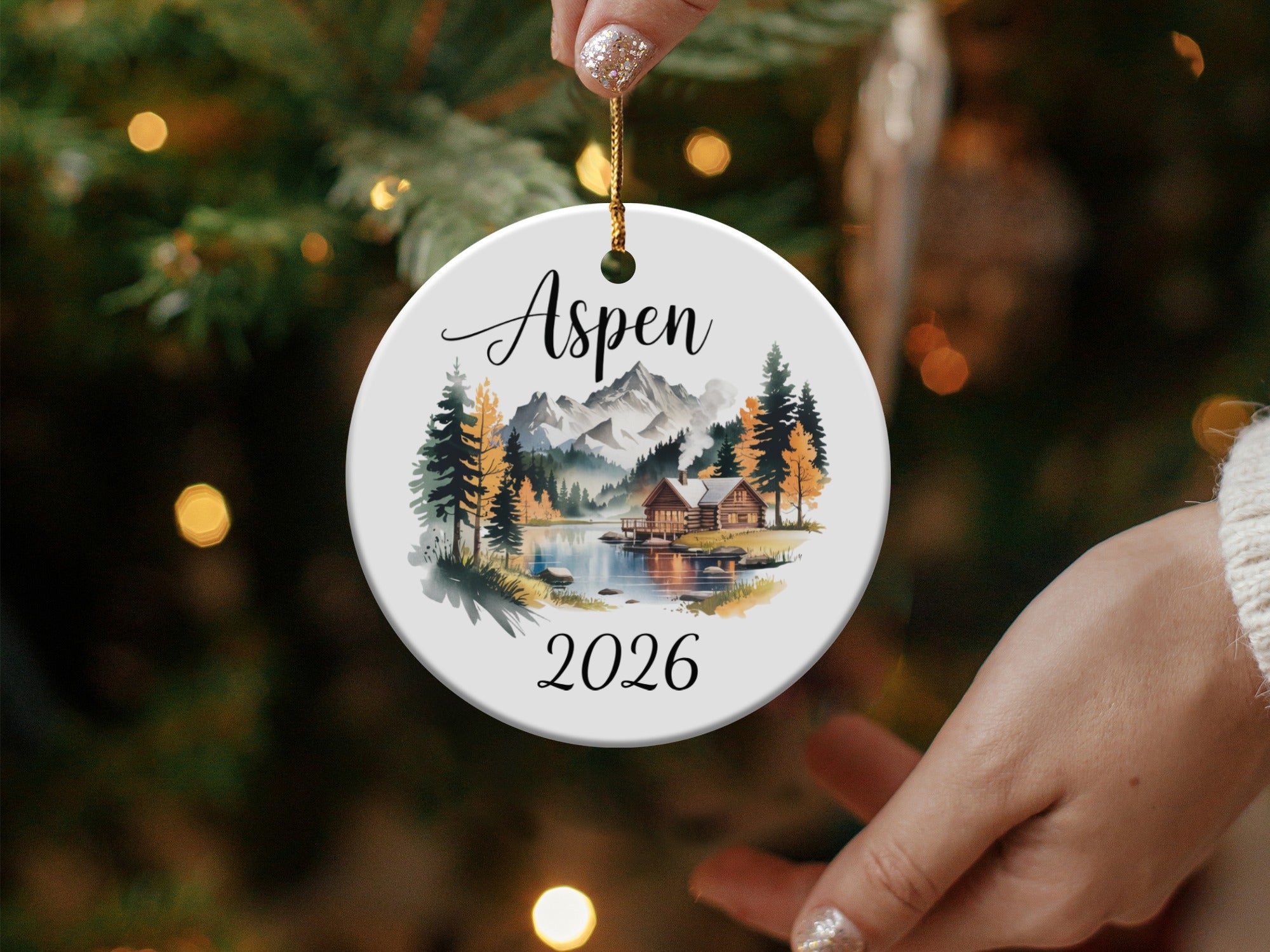 Round ceramic ornament with "Aspen 2026" and a mountain cabin scene.
