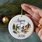 Round ornament with Aspen 2026 text and a cabin in a forest scene.
