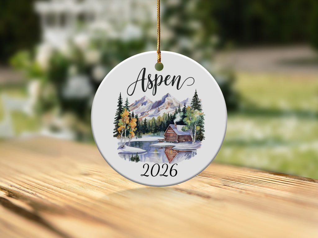 Round ornament with a cabin by a lake and mountains, reading "Aspen 2026".