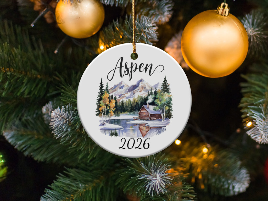 Circular ornament with "Aspen 2026" above a cabin scene and snow-capped mountains.