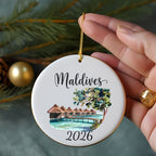 Round ornament with "Maldives" and "2026" above a watercolor scene of huts on stilts.