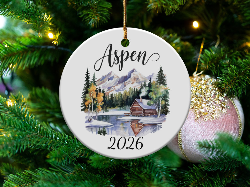 Round ornament with "Aspen 2026" and a log cabin scene.